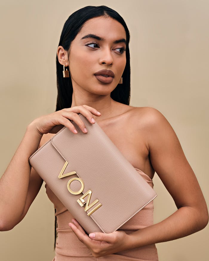 Portrait of a woman holding a beige designer clutch with gold lettering against a neutral background.